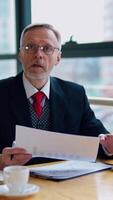 Businessman is checking a printed report or document while sitting at table, working on paper work at a laptop explaining his point of view. in an office. Vertical video