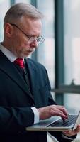Serious businessman with laptop in hands thinking about future meeting checking presentation standing near window with panoramic city view. from the side. Vertical video
