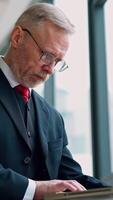 Senior serious businessman standing near window with panoramic city view. Laptop in hands, man thinking while checking e-mails standing near window. from the side. Vertical video