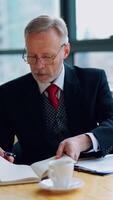 Smart mature businessman working in the office. Senior man in elegant suit sitting at the desk and using notebook in his work. Business concept. Vertical video