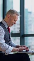 Happy mature man is typing on a laptop near the window. Entrepreneur in suit sitting near the window and working in the office on the city background. Vertical video
