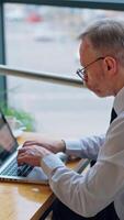 Side view of businessman working on a computer. Mature man is typing on the laptop in the office on the city background through the window. Vertical video