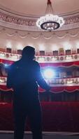 ator em etapa dentro Holofote. ativo homem em pé em etapa do uma clássico teatro com linhas do vermelho cadeiras sem público. costas visualizar. vertical video