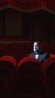 Rich man among rows of empty red chairs. Businessman in medical mask sitting in comfortable chairs alone on dark theater background. Pandemic in the world. Vertical video