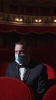 Alone businessman in mask in theater. Man in protective face mask sitting in a chair of empty auditorium. Theater in quarantine. Coronavirus pandemic. Vertical video