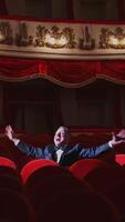 Successful man in suit alone in large auditorium. Actor man is sitting among empty red armchairs and singing in opera house. Vertical video