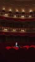 Spectator in mask sitting alone in empty theater. Man putting off his facial mask and talking in large dark auditorium among many rows of vacant red chairs in the theater. Vertical video