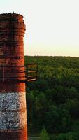 Brick industrial tower on nature background. Old pipe in ruined plant at sunset. Chimney surrounded by green trees and river in the countryside. Aerial view. Vertical video