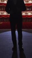 Man in suit standing on stage back to auditorium. Dark silhouette of unrecognizable actor on the background of classic theatre hall in red and white colors. Vertical video