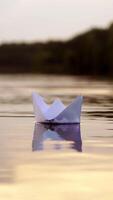 White paper boat is floating on the water near the forest background in the evening. Origami boat floats away into the distance along the river Vertical video