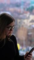 Beautiful female with cup of coffee thoughtfully browsing smartphone while standing by the window Vertical video