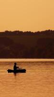 Silhouette of a man with fishing rods in the middle of the lake at sunset. Natural river background in the evening and a fisherman in a boat. Vertical video