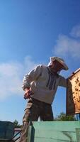 Man in protective hat on apiary. Beekeeper works with wooden frame from hive under blue sky. View from below. Apiculture. Vertical video