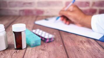A doctor's hand writing on a notepad with a pen and pills on a wooden table video