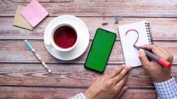 Man writing question mark on notebook with smartphone and cup of coffee on wooden table video