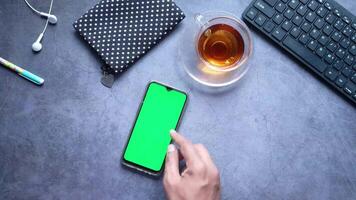 Hand holding a smartphone with a green screen and a cup of tea on a desk video