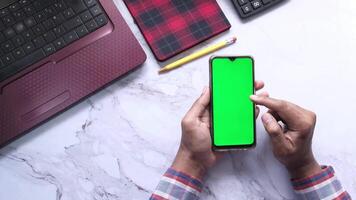 Hands holding a smartphone with a green screen on a desk with a laptop and a notebook video