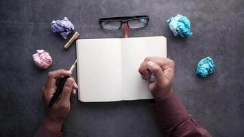 A man's hands writing in a notebook with crumpled paper and a pen on a dark video