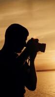 A man with a camera in his hands is photographing the sunset by the river on the background of people bathing. Close-up Vertical video