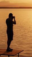 un talentoso fotógrafo con un cámara en su manos tomando imágenes de el puesta de sol en el río en el puente con un banco. silueta de hombre con un cámara en su favorito trabajar. vertical video