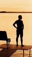 A man was standing on the bridge and than sat down on a bench to looking at the beauty of nature on the background of the river and the sunset on a summer evening. Vertical video