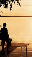 A young man goes to the bridge on the river and looks on a beautiful view of the sunset. Orange background. Vertical video
