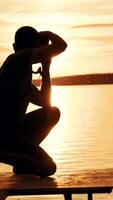 A man in shorts and a t-shirt is squatting and holding a camera focused on the sunset against the background of the forest and people in the river. Vertical video