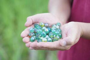 Close up hands hold round small marble balls, spherical objects often made from glass. Green background. Concept, object use as toy for playing games or use for DIY craft decoration. photo