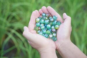 Close up hands hold round small marble balls, spherical objects often made from glass. Green background. Concept, object use as toy for playing games or use for DIY craft decoration. photo
