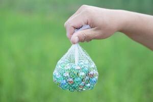Hand holds net bag of marbles balls small spherical object often made from glass. Green background. Concept. object use as toy for playing games or use for DIY craft decoration. photo