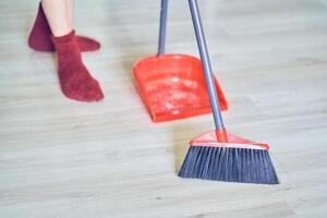 Close up view of a red broom and plastic dustpan on the floor with person's legs in red socks. Worker, cleaner using cleaning items with copy space. Concept of cleaning dust, home and sweeping floor photo