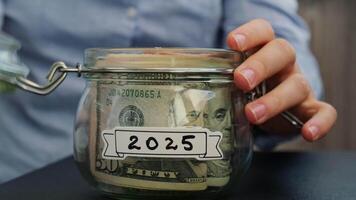 Unrecognizable woman add money to Saving Money In Glass Jar filled with Dollars banknotes. 2025 year transcription in front of jar. Managing personal finances extra income for future insecurity background video