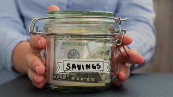 Female hands put on table Saving Money In Glass Jar filled with Dollars banknotes. DONATE transcription in front of jar. Managing personal finances extra income for future insecurity background video