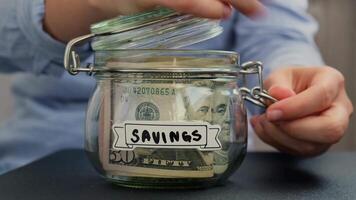 Unrecognizable woman add money to Saving Money In Glass Jar filled with Dollars banknotes. SAVINGS transcription in front of jar. Managing personal finances extra income for future insecurity background video