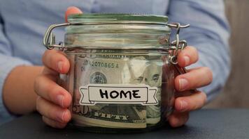 Unrecognizable woman put on table Saving Money In Glass Jar filled with Dollars banknotes. HOME transcription in front of jar. Managing personal finances extra income for future insecurity background video