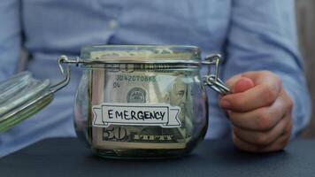Female hands add money to Saving Money In Glass Jar filled with Dollars banknotes. EMERGENCY transcription in front of jar. Managing personal finances extra income for future insecurity background video