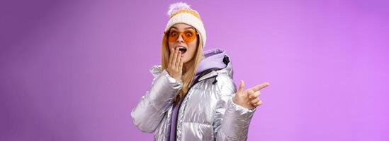 Amazed speechless surprised attractive stylish blond girlfriend in sunglasses, silver glittering jacket hat drop jaw impressed widen eyes shocked pointing left astonished, purple background photo