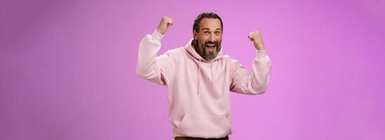 Yeah who cares age. Carefree delighted happy old man bearded in trendy pink hoodie raise fists joyfully triumphing having fun glad win celebrating success accomplish goal, posing purple background photo