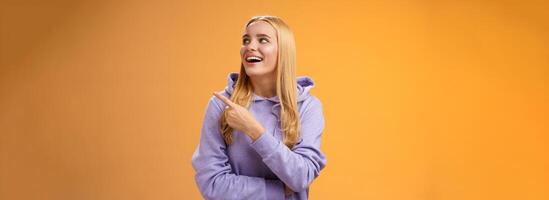 Amused carefree joyful blond european woman in hoodie pointing looking right pleased smiling happily enjoying interesting fascinating peromance making choice in store shopping, orange background photo