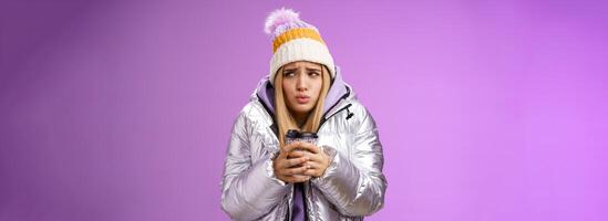 Trembling uncomfortable cute pouting young blond girl feel freezing cold winter snowy weather outside shaking low temperature hold take-away hot coffee cup warming up, purple background photo