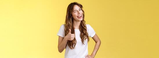 I support you guys good luck. Happy cheerful cheeky attractive girl wink approval like show thumb up smiling broadly satisfied awesome party congratulating friend well done yellow background photo