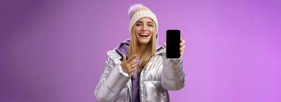 Satisfied amused good-looking blond girl suggest take look smartphone display smiling happily pointing mobile phone delighted talking about awesome new app features, standing purple background photo