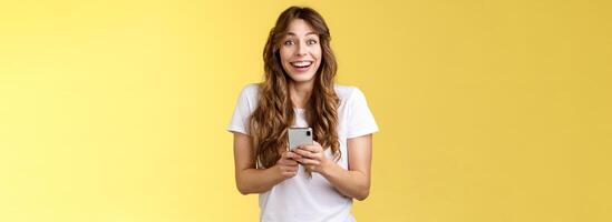 Extremely happy enthusiastic surprised joyful lovely feminine girl receive pleasant touching message online hold smartphone smiling broadly look camera joyful amused yellow background photo