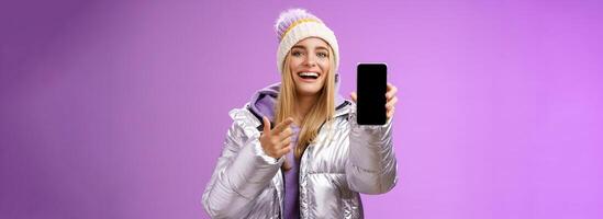 Satisfied amused good-looking blond girl suggest take look smartphone display smiling happily pointing mobile phone delighted talking about awesome new app features, standing purple background photo