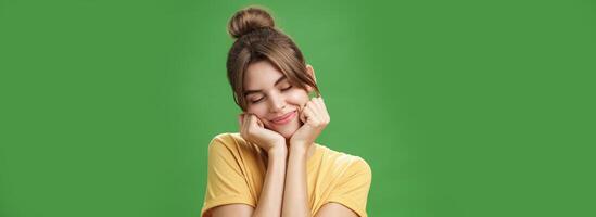 Tender sensual timid european girl in yellow t-shirt leaning head on shoulder touching cheeks with hands closing eyes and smiling with soft grin feeling nostalgic and romantic over green background photo