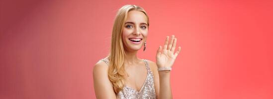 Waist-up studio shot friendly attractive elegant tender blond woman say hi waving raised hand greeting welcoming friend smiling delighted introducing herself hello gesture, red background photo
