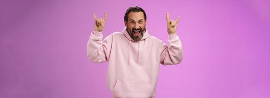 Carefree enthusiastic excited mature bearded man grey hair having fun show rock-n-roll gesture attend awesome heavy metal concert grimacing thrilled looking happy upbeat, purple background photo