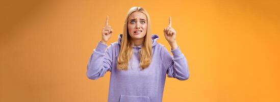 Worried perplexed unsure cute blond woman presenting project standing awkward afraid humiliation crining scared pointing up raised index finger waiting reply nervously, orange background photo