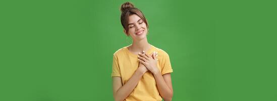 Happy kind and timid young attractive woman with gapped teeth smiling sincere and grateful closing eyes pressing hands on chest in thankful pose, daydreaming, recalling nice tender memories photo