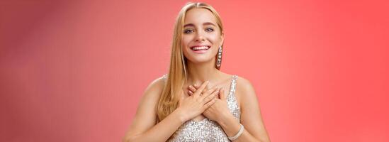 Grateful charming blond european 25s woman in silver party dress press palms heart feel thankful appreciate effort cherish romantic gesture receive flattering compliments gifts, smiling happily photo